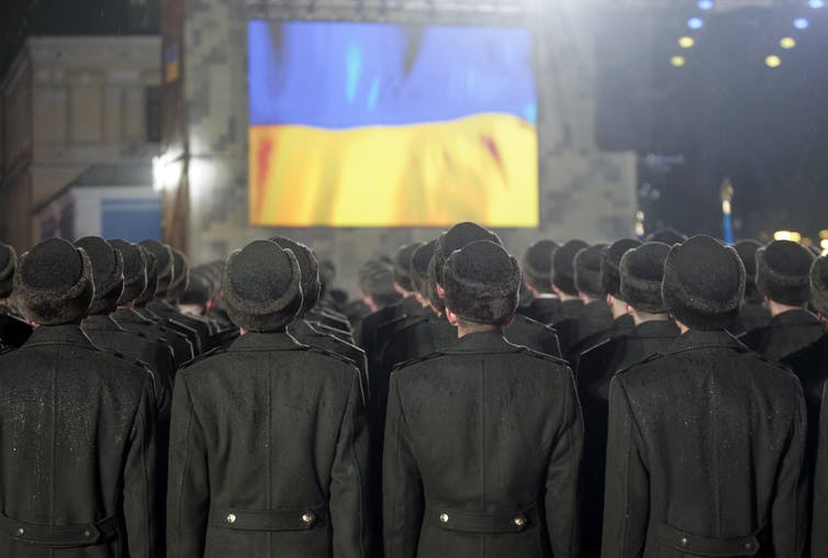 Soldados permanecen en fila, de espaldas a la cámara, frente a una bandera ucraniana.