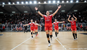 Female handball players in a sports hall during a match, one player in red uniform celebrating a goal while teammates chee...