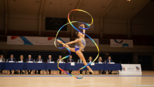 Young female gymnast performing rhythmic gymnastics routine in a sports pavilion, colorful ribbon flowing through the air,...