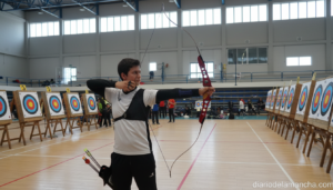 Archer in indoor sports pavilion, aiming recurve bow and arrow at target during a local competition, colorful circular tar...