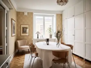 Comedor con papel pintado dorado botánico, mesa ovalada con mantel blanco y sillas de madera con patas metálicas