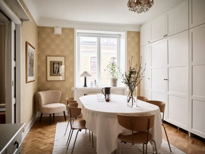 Comedor con papel pintado dorado botánico, mesa ovalada con mantel blanco y sillas de madera con patas metálicas
