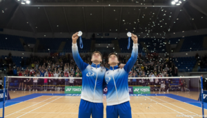 Two badminton players celebrating with silver medals at indoor sports court after competition, fans in background, bright ...