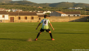 A young football defender in a white and green jersey training on a grass pitch in a small Spanish town stadium, action sh...