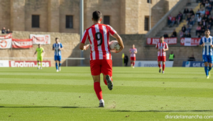 A football player in a red and white jersey running with the ball on a green grass pitch during a regional Spanish footbal...