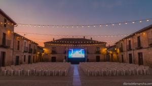 Wide shot of a small Spanish village square at dusk with a large outdoor LED screen set up for a public football viewing e...