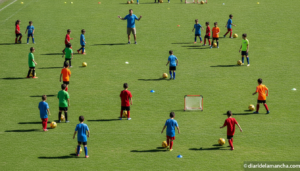 Wide realistic photo of a children's summer football camp on a municipal outdoor sports pitch in Spain, kids in colorful s...
