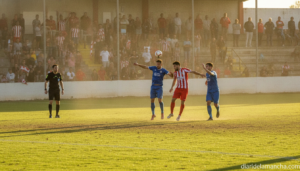 Football match in a Spanish lower-league stadium, players competing for a header near the goal area, referee watching, mod...